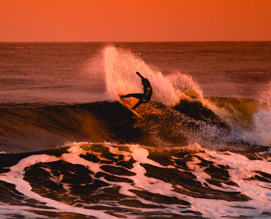 A surfer doing a maneuver on a wave during sunset